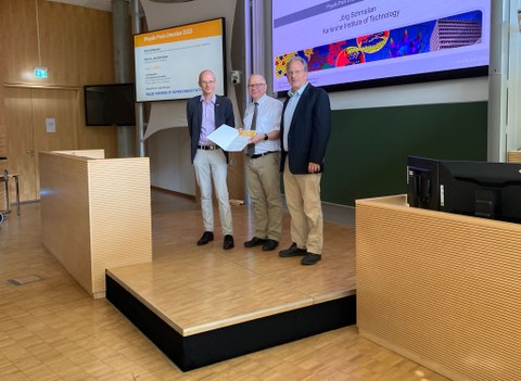 Drei Männer stehen auf einer Bühne. Im Hintergrund wird eine Präsentationsfolie zum Physik-Preis Dresden 2023 angezeigt. Der Mann in der Mitte hält eine Urkunde in der Hand.
