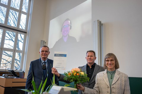 Drei Personen stehen im Vordergrund mit einer Urkunde und Blumen. Im Hintergrund ist eine Person an der Leinwand per Video zugeschalten.