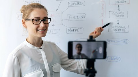 Das Foto zeigt eine junge Frau vor einem Whiteboard. Sie spricht in ihr Handy, welches die Frau filmt.