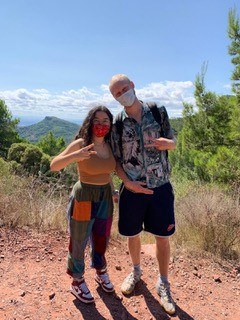 Zwei Personen mit Mund-Nasenbedeckung stehen in einer malerischen Landschaft und schauen in die Kamera.