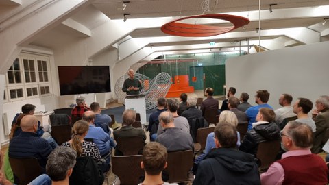 Ein Vortragender steht vor Publikum im Erlebnisland Mathematik. Im Hintergrund sieht man den sogenannten "Krabbelknoten".