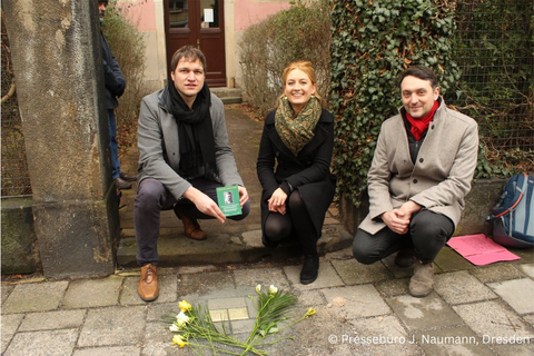 Drei Menschen knieen hinter den Stolpersteinen des Ehepaars Blumenfeld. um die Steine sind Schnittblumen gelegt. Die drei Menschen, die für das Bild posieren, sind zwei Männer und eine Frau in der Mitte