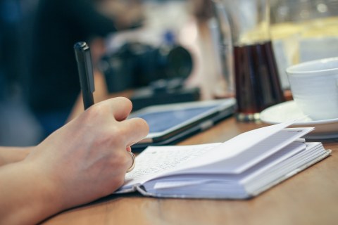 ein Schreibtisch, im Vordergrund eine Hand mit Stift