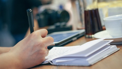 ein Schreibtisch, im Vordergrund eine Hand mit Stift