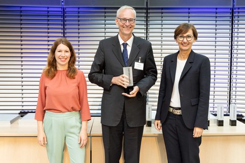 zwischen zwei Frauen steht ein Mann mit einem Pokal in der Hand. Alle drei lachen in die Kamera.