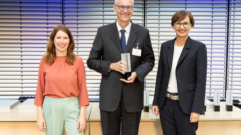 zwischen zwei Frauen steht ein Mann mit einem Pokal in der Hand. Alle drei lachen in die Kamera.