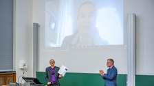 Zwei Professoren stehen vorn im Hörsaal. An der Wand ist eine Videoschalte projeziert.