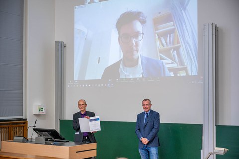 Der Dekan zeigt die Urkunde, während im Hintergrund der Preisträger in einer Videoschalte gezeigt wird.