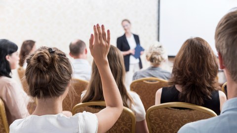 Das Foto zeigt eine Gruppe von Menschen, die sich die Präsentation einer Person anhört. Eine Frau im Vordergrund des Bildes hebt ihren Arm für eine Wortmeldung.