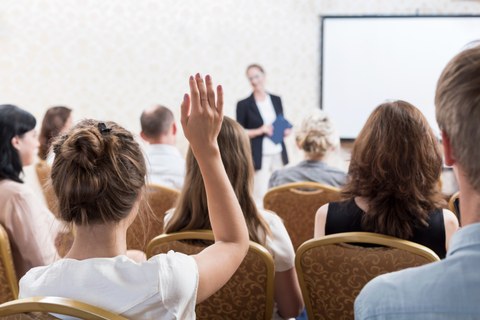 Das Foto zeigt eine Gruppe von Menschen, die sich die Präsentation einer Person anhört. Eine Frau im Vordergrund des Bildes hebt ihren Arm für eine Wortmeldung.