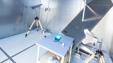 Das Foto zeigt einen Raum mit metallischen Wänden. In dem Raum stehen mehrere Geräte zum Messen von elektrischer Strahlung. In der Mitte steht ein Tisch mit einem kleinen, grün leuchtendem Gerät.