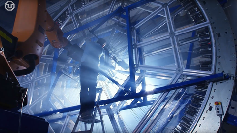 Eine Person steht auf einer Leiter in einer großen Faserwickelmaschine. Um sie verlaufen viele gespannte Fasern. Links ist ein Industrieroboterarm zu sehen. Lichtstrahlen und Nebel erzeugen eine futuristische Atmosphäre.