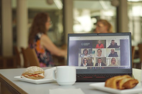 Ein Laptop , Geschirr und Schreibmaterial  auf einem Tisch, zwei Menschen unscharf im Hintergrund beim Gespräch