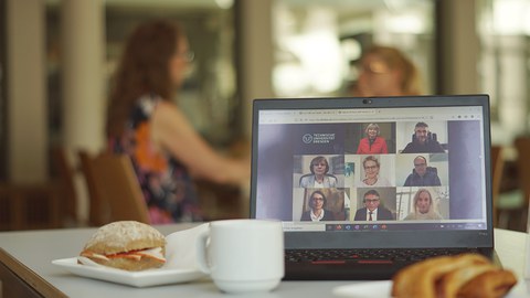 Ein Laptop , Geschirr und Schreibmaterial  auf einem Tisch, zwei Menschen unscharf im Hintergrund beim Gespräch