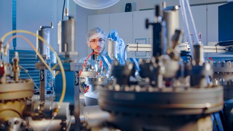 Foto: Eine Laborumgebung mit verschiedenen wissenschaftlichen Geräten im Vordergrund, darunter runde Maschinen und Rohre. Im Hintergrund ist eine Person in Schutzkleidung zu sehen.