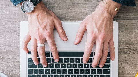 Das Foto zeigt von oben die Hände einer Person, welche eine Tastatur bedienen, daneben liegen zur linken Seite Kopfhörer und zur rechten ein gelbes Buch.