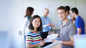 Das Foto zeigt zwei Studierende. Sie stehen sich gegenüber, schauen in die Kamera und halten gemeinsam mehrere Blätter in den Händen. Im Hintergrund erkennt man weitere Personen vor einem Whiteboard. 