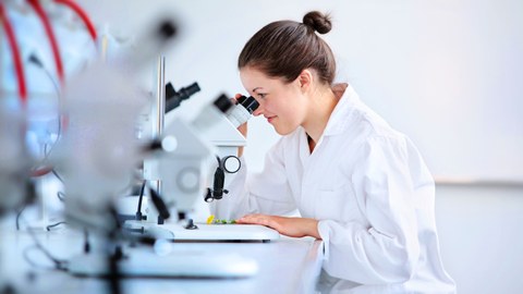 Das Foto zeigt eine Studentin mit Laborkittel. Sie sitzt an einem Tisch und schaut durch ein Mikroskop.