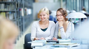 Das Foto zeigt zwei Studentinnen in der Bibliothek. Sie sitzen an einem Tisch und lächeln in die Kamera. Vor ihnen liegen viele Bücher. 