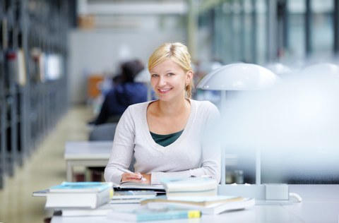 Das Foto zeigt eine Studentin in der Bibliothek. Sie sitzt an einem Tisch und lächelt in die Kamera. Vor ihr liegen viele Bücher. 