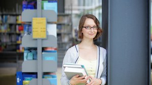  Das Foto zeigt eine Studentin in der Bibliothek. Sie steht an einem Pfeiler, hält einige Bücher in den Händen und lächelt in die Kamera.