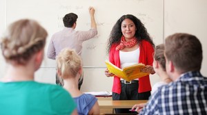  Das Foto zeigt ein Fremdsprachenseminar aus Sicht eines Studierenden. Vor der Tafel im Hintergrund steht die Dozentin. Sie hält ein aufgeschlagenes Buch in den Händen. An der Tafel steht ein Student, der gerade mit einem Stift etwas anschreibt. 