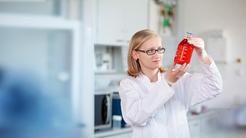  Das Foto zeigt eine Frau mit weißem Laborkittel in einem Labor. In der Hand hält sie ein Glasgefäß mit einer roten Flüssigkeit, welches sie intensiv betrachtet. 