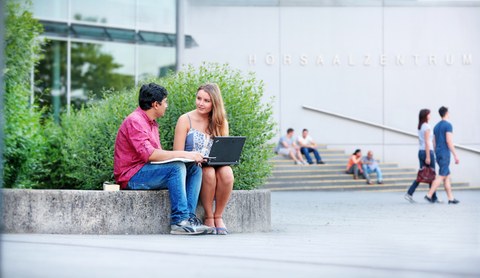  Das Foto zeigt zwei Studierende vor dem Hörsaalzentrum der TU Dresden. Sie sitzen auf einer steinernen Abgrenzung und reden miteinander. Während die Frau einen Laptop auf dem Schoß hat, hält der Mann ein Buch in den Händen.