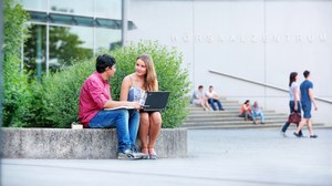  Das Foto zeigt zwei Studierende vor dem Hörsaalzentrum der TU Dresden. Sie sitzen auf einer steinernen Abgrenzung und reden miteinander. Während die Frau einen Laptop auf dem Schoß hat, hält der Mann ein Buch in den Händen.