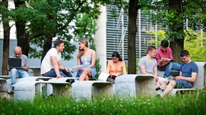  Das Foto zeigt mehrere Studierende draußen im Grünen. Sie sitzen auf einer geschwungenen Betonformation aus Tischen und Bänken und lernen. 