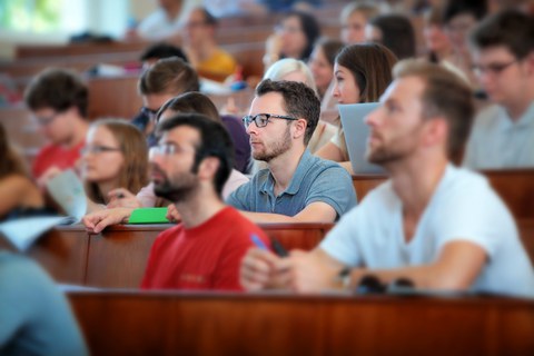  Das Foto zeigt einen Hörsaal voller Studierender. Die Studierenden schauen nach vorn und hören dem Referenten aufmerksam zu. Der Fokus des Bildes liegt lediglich auf einer Person in der Mitte. 