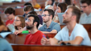  Das Foto zeigt einen Hörsaal voller Studierender. Die Studierenden schauen nach vorn und hören dem Referenten aufmerksam zu. Der Fokus des Bildes liegt lediglich auf einer Person in der Mitte. 