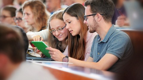  Das Foto zeigt drei Studierende in einem vollen Hörsaal. Sie sitzen nebeneinander und schauen gemeinsam in ein Notebook.