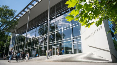 Außenansicht des Hörsaalzentrums mit großer Glasfassade, Treppen und mehreren Personen auf dem Vorplatz unter blauem Himmel.