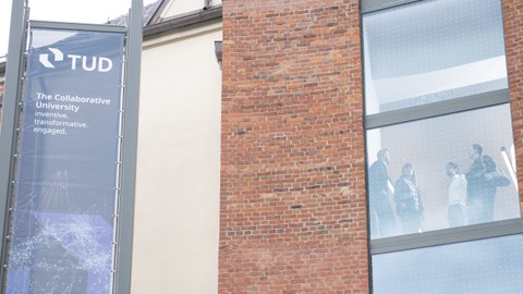 Banner mit dem Logo der TUD und dem Text 'The Collaborative University. Transfervative. Transformative. Engaged.' neben einem Backsteingebäude mit großen Fenstern, hinter denen mehrere Personen stehen.
