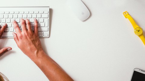 Das Foto zeigt einen weißen Schreibtisch von oben, an welchem eine Person auf einer Tastatur tippt. Auf dem Tisch liegen außerdem eine weiße Computer-Maus, ein Smartphone und eine gelbe Armbanduhr.