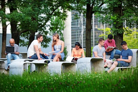  Das Foto zeigt mehrere Studierende draußen im Grünen. Sie sitzen auf einer geschwungenen Betonformation aus Tischen und Bänken und lernen. 