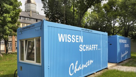 Das Foto zeigt zwei blaue Container mit der Aufschrift "Wissen schafft... Chancen" und "Knowledge creates... fascination", welche auf einer Wiese des Campus der TU Dresden stehen.
