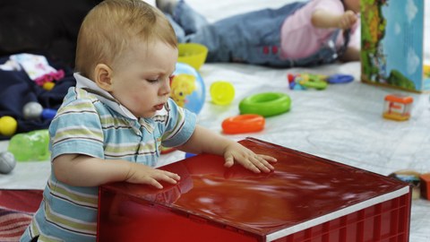 Das Foto zeigt ein Kleinkind, dass mit einer großen roten Kiste spielt. Im Hintergrund blättert ein anderes Kleinkind in einem Buch. 