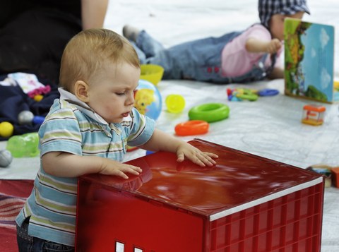 Das Foto zeigt ein Kleinkind, dass mit einer großen roten Kiste spielt. Im Hintergrund blättert ein anderes Kleinkind in einem Buch. 