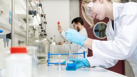 Foto zeigt zwei Personen, die im Labor arbeiten. Sie tragen Arbeitskleidung und Schutzbrille.