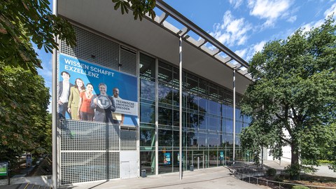 Frontansicht des Hörsaalzentrums der TU Dresden. Am Gebäude Hängt ein Poster mit der Aufschrift: "Wissen Schafft Exzellenz".