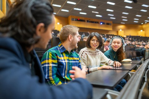 Fotos von vier sich unterhaltenden Studierenden in einem vollen TUD-Hörsaal.