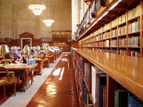 Das Foto zeigt den Lesesaal einer Bibliothek. Auf der linken Bildhälfte sind Arbeitsplätze, an welchen Studierende sitzen. Die recht Bildhälfte zeigt eine lange Reihe von Bücherregalen.