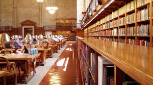 Das Foto zeigt den Lesesaal einer Bibliothek. Auf der linken Bildhälfte sind Arbeitsplätze, an welchen Studierende sitzen. Die recht Bildhälfte zeigt eine lange Reihe von Bücherregalen.
