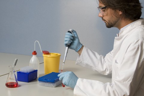 Das Foto zeigt eine Person, welche Arbeitskleidung und Schutzbrille tragend an einem Tisch sitzt, auf welchem verschiedene Laborutensilien stehen. Die Person füllt mit einer Spritze eine rote Flüssigkeit in ein Reagenzglas. 