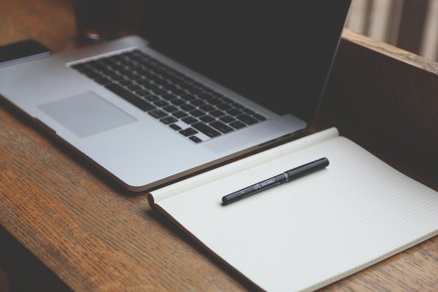 Das Foto zeigt einen Laptop mit schwarzem Bildschirm, rechts daneben ein geöffnetes, leeres Notizbuch und einen daraufliegenden Stift  auf einer hölzernen Oberfläche.
