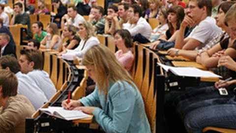  Das Foto zeigt einen Hörsaal voller Studierender während einer Vorlesung. Viele der Studierenden schauen nach vorn oder machen sich Notizen.