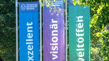 Foto von drei im Wind wehenden Flaggen vor dem Rektoratsgebäude in blau, lila und grün. Darauf steht "exzellent", "visionär" und "weltoffen".