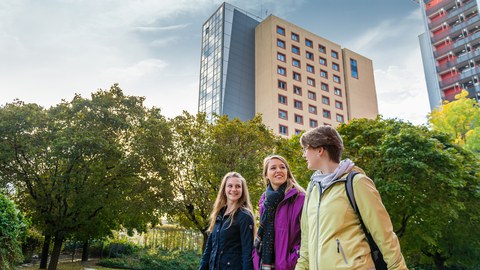Das Foto zeigt drei Studentinnen. Sie laufen gemeinsam durch einen Park und reden miteinander. Im Hintergrund erkennt man Studentenwohnheime.